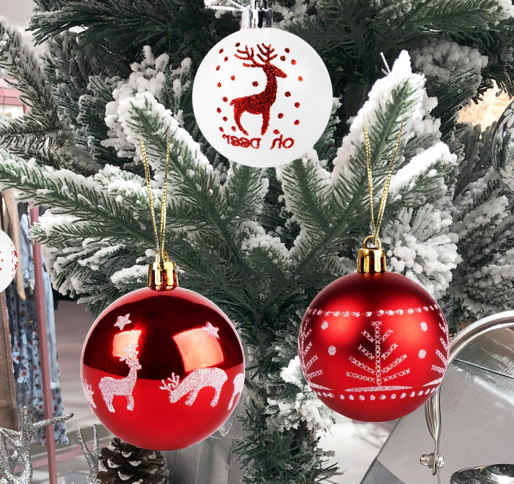 Three shatterproof Christmas ball ornaments hanging on a flocked tree branch, one white with a red reindeer and text, two red with white glitter patterns.