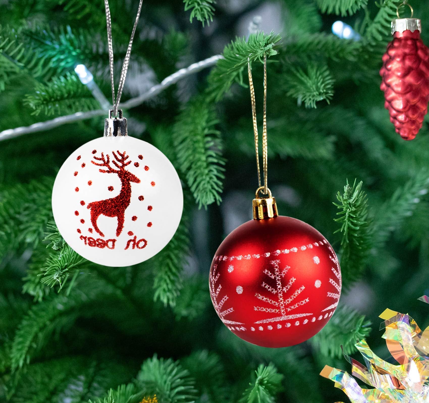 Two Christmas ball ornaments, one white with a red glitter reindeer and dots, the other red with white snowflake patterns, hanging on a Christmas tree.