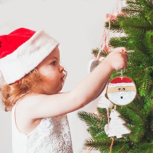 Young child wearing a Santa hat hangs a wooden Santa ornament on a Christmas tree