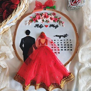 Embroidery hoop art with bride and groom figures, floral accents, and a calendar marking August, set against a white fabric background.
