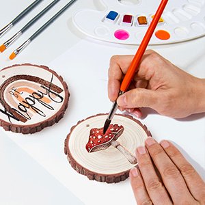 Hand painting a mushroom on a natural wood slice ornament with holes and twine, watercolor palette and brushes nearby.