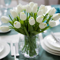 A vase of white artificial tulips arranged on a table setting with plates and cutlery.