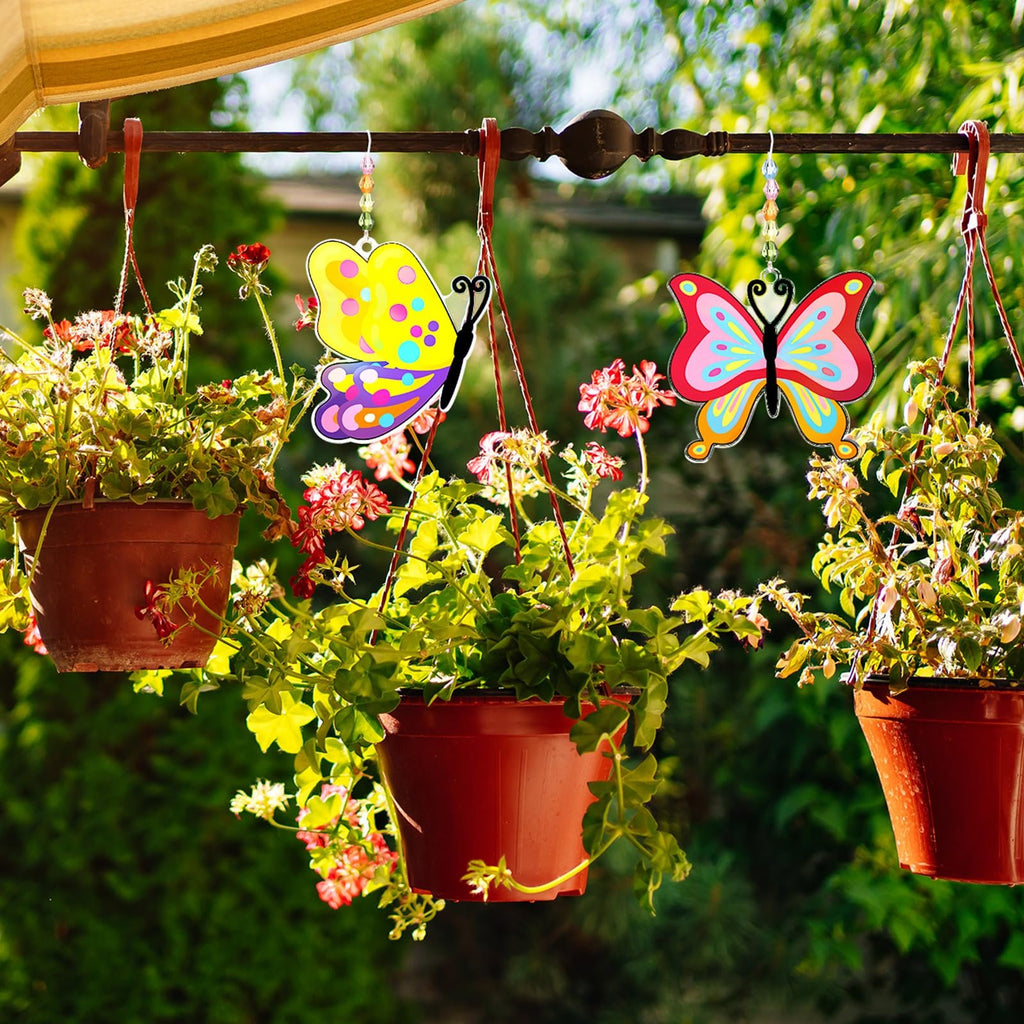 Two colorful butterfly suncatchers hanging outdoors near potted plants, one yellow and purple, the other pink and blue.
