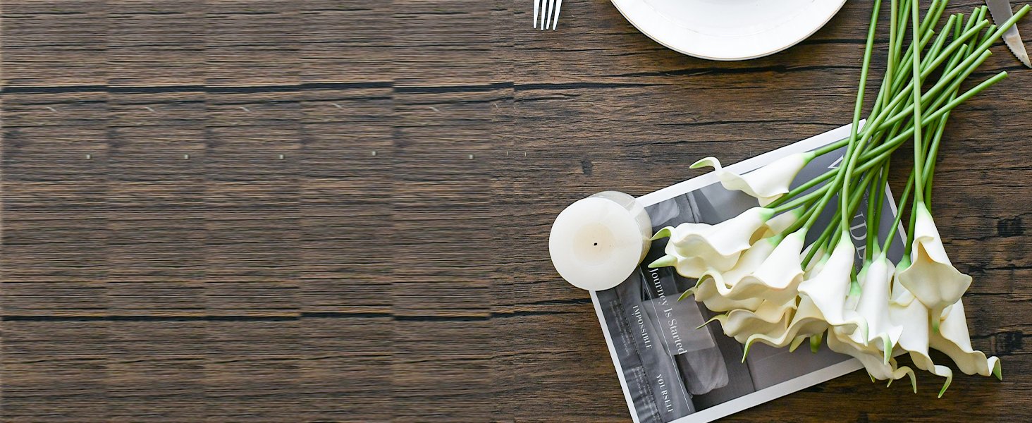 A bouquet of white calla lily artificial flowers arranged on a wooden table with a candle and a magazine.