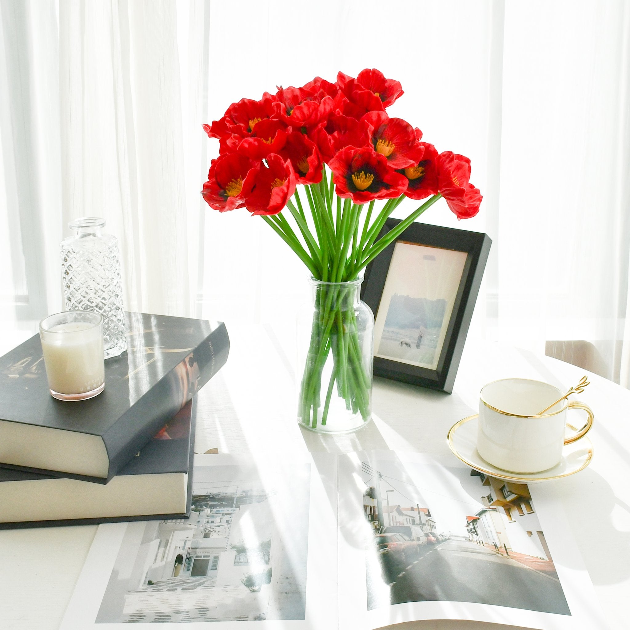 A bouquet of 30 red artificial poppies in a clear glass vase on a table with books, a candle, and a teacup.
