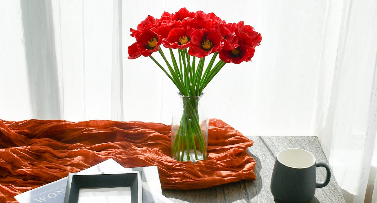 A bouquet of 30 red artificial poppies in a clear glass vase on an orange textured runner, with a dark mug and picture frame nearby.