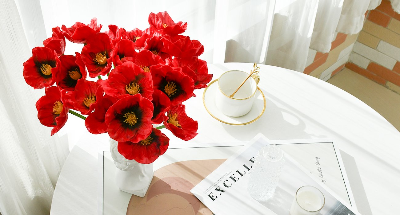 A bouquet of 30 red artificial poppies in a white bust vase on a white table with a teacup and saucer, a glass vase, and a magazine.