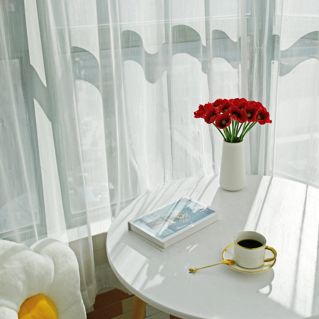 A vase of red artificial poppies sits on a white table next to a book and a cup of coffee.