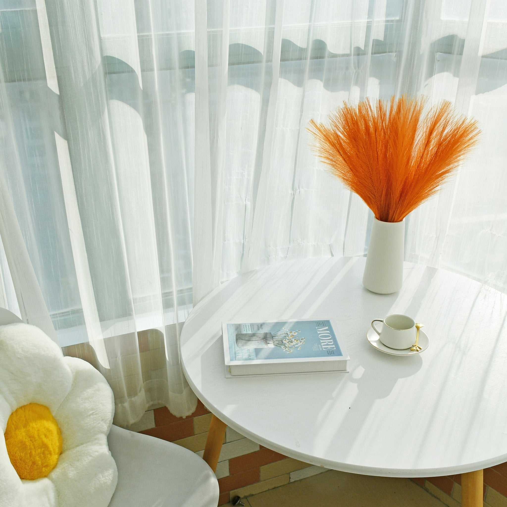 White table with a vase of orange pampas grass, a book, and a coffee cup with a gold spoon