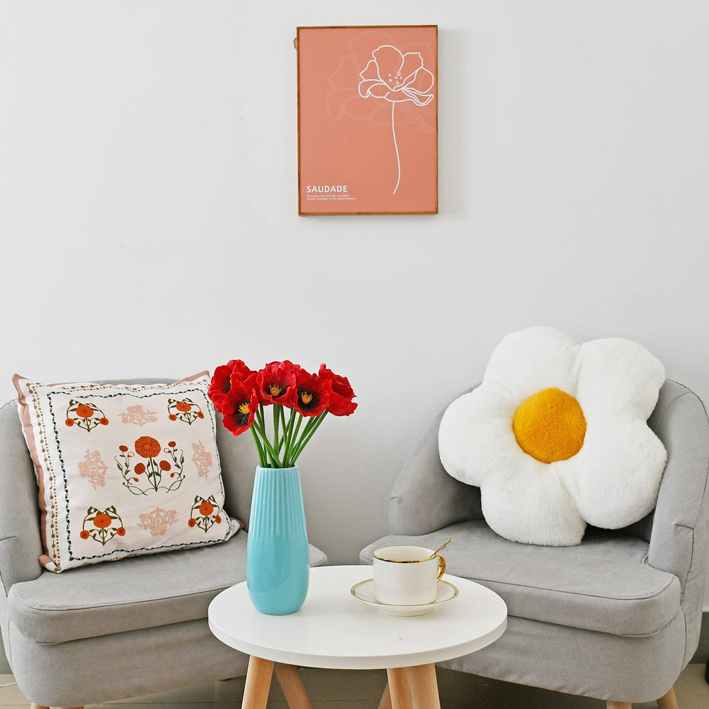 Red artificial poppies in a blue vase on a white table between two chairs, with a decorative pillow and a framed artwork.