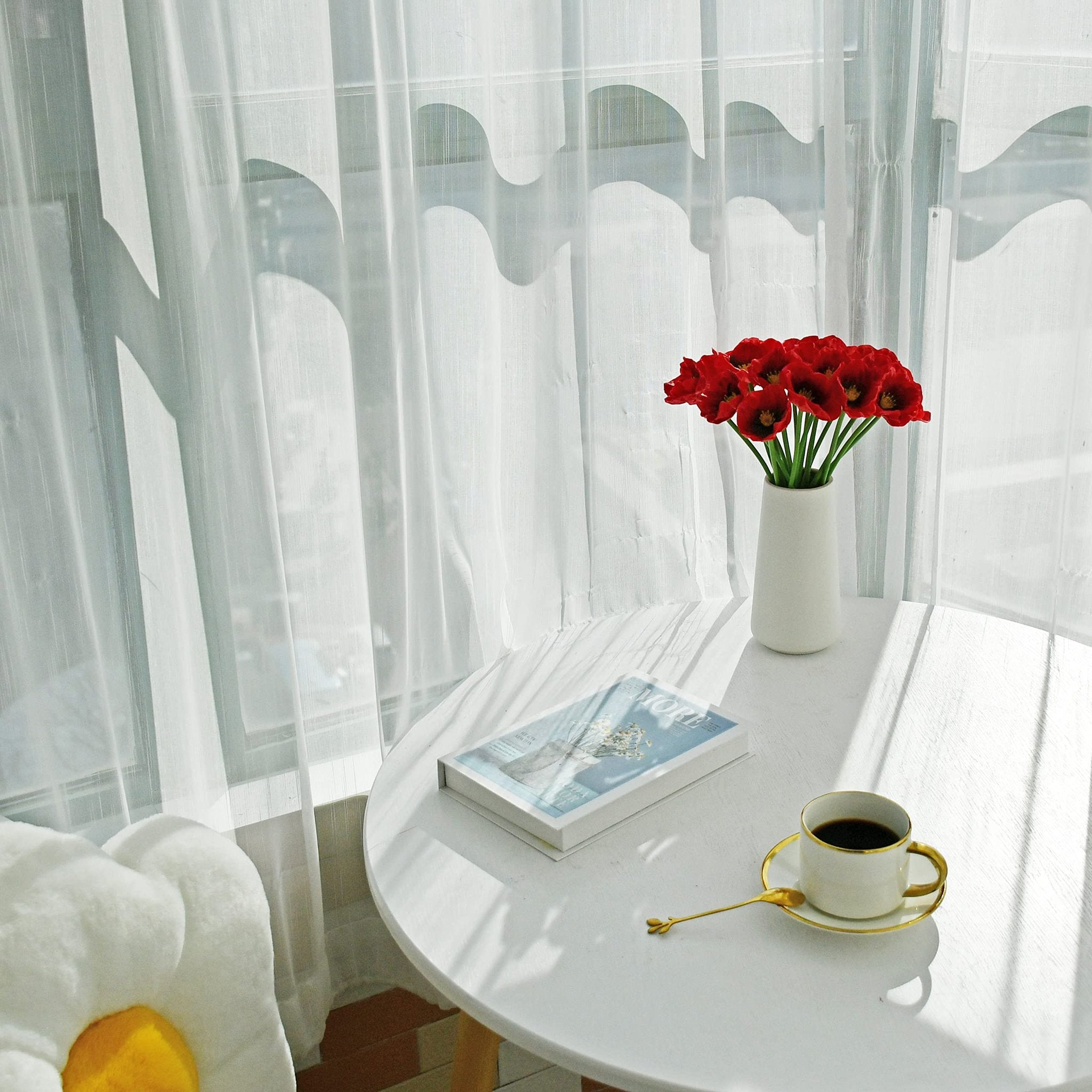 A vase of red artificial poppies sits on a white table next to a book and a cup of coffee.