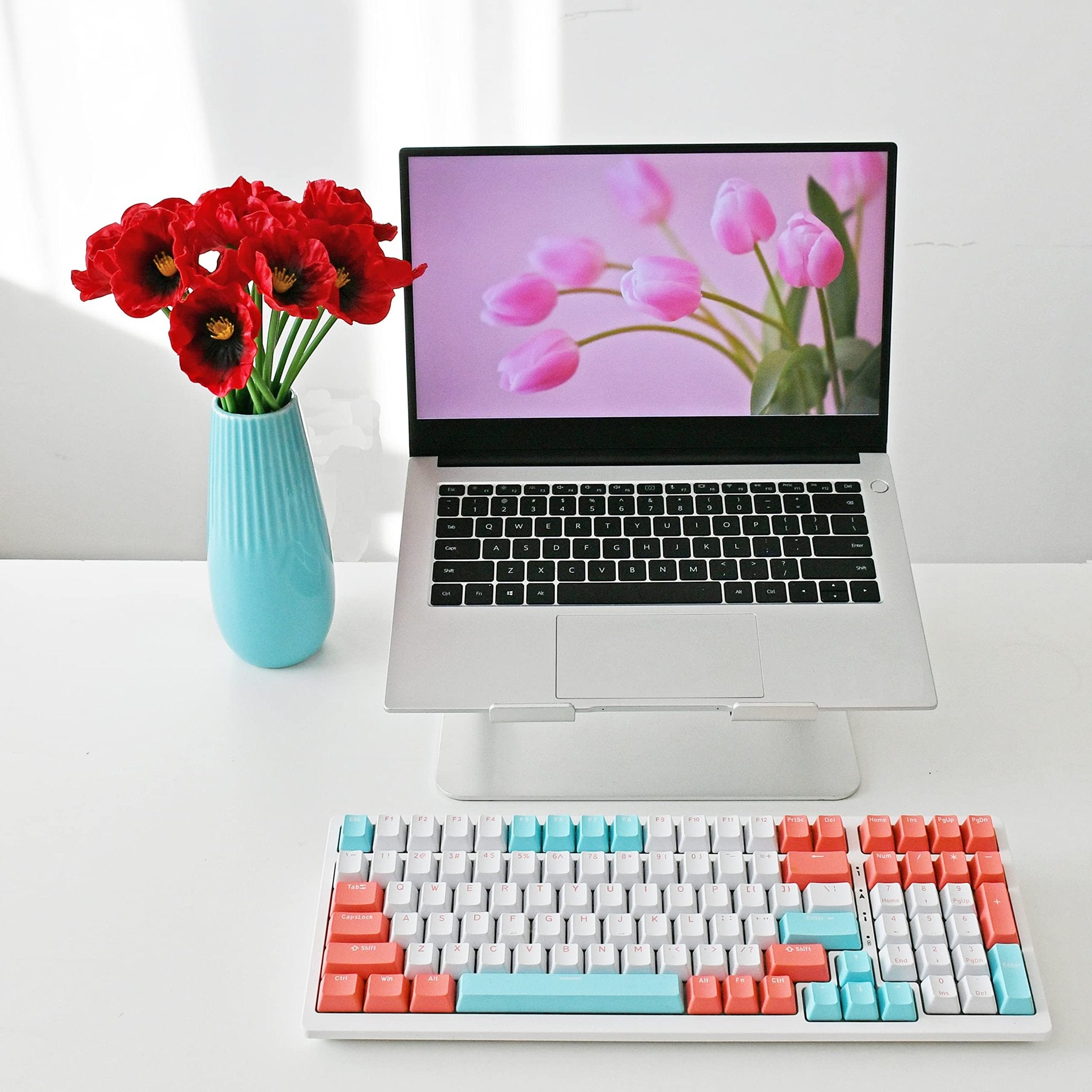 A vase of red artificial poppies sits next to a laptop displaying pink tulips on its screen, with a colorful keyboard in the foreground.