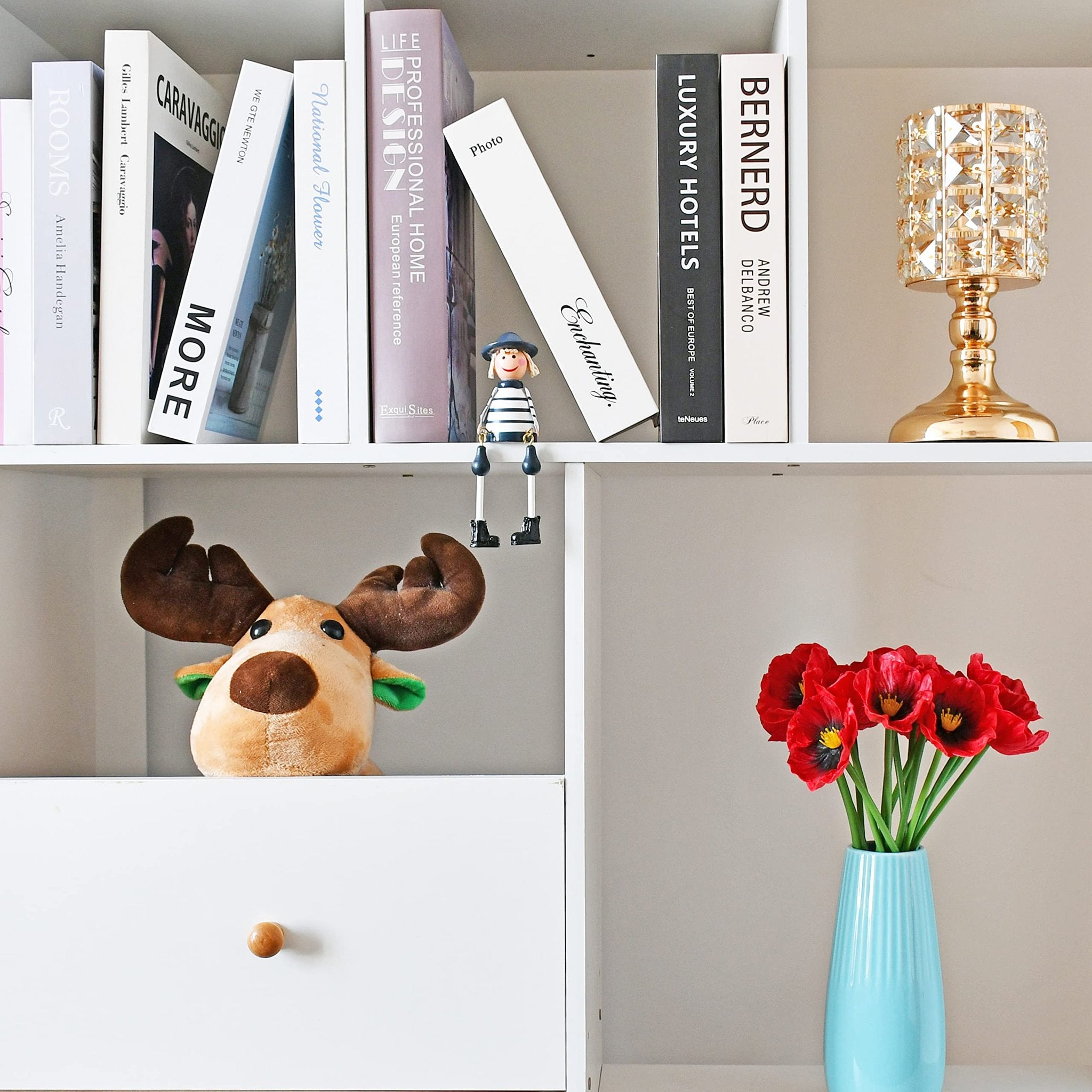 A bouquet of red artificial poppies in a light blue vase on a shelf with books and a decorative lamp.