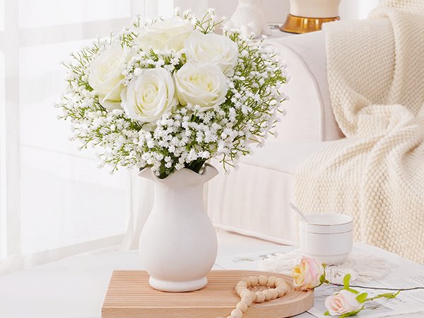 White roses and baby's breath flowers in a white vase on a wooden tray, with a knitted blanket in the background.