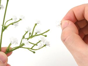 Hand holding a single artificial baby's breath flower blossom next to a stem with multiple white blossoms against a white background.