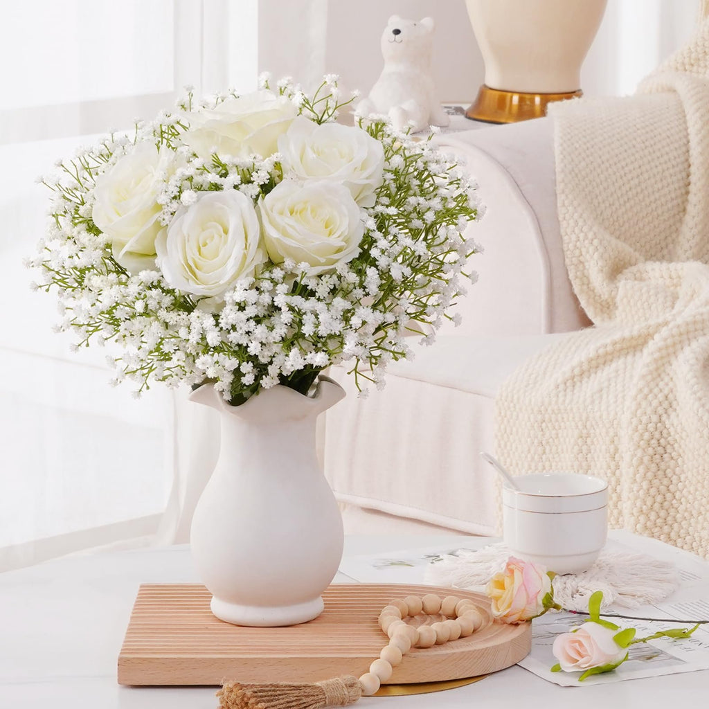 White roses and baby's breath flowers in a white vase on a wooden tray with decorative beads and a cup.