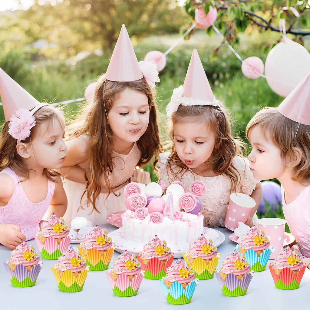 Four young girls in party hats blowing out candles on a birthday cake, with decorated cupcakes in the foreground.