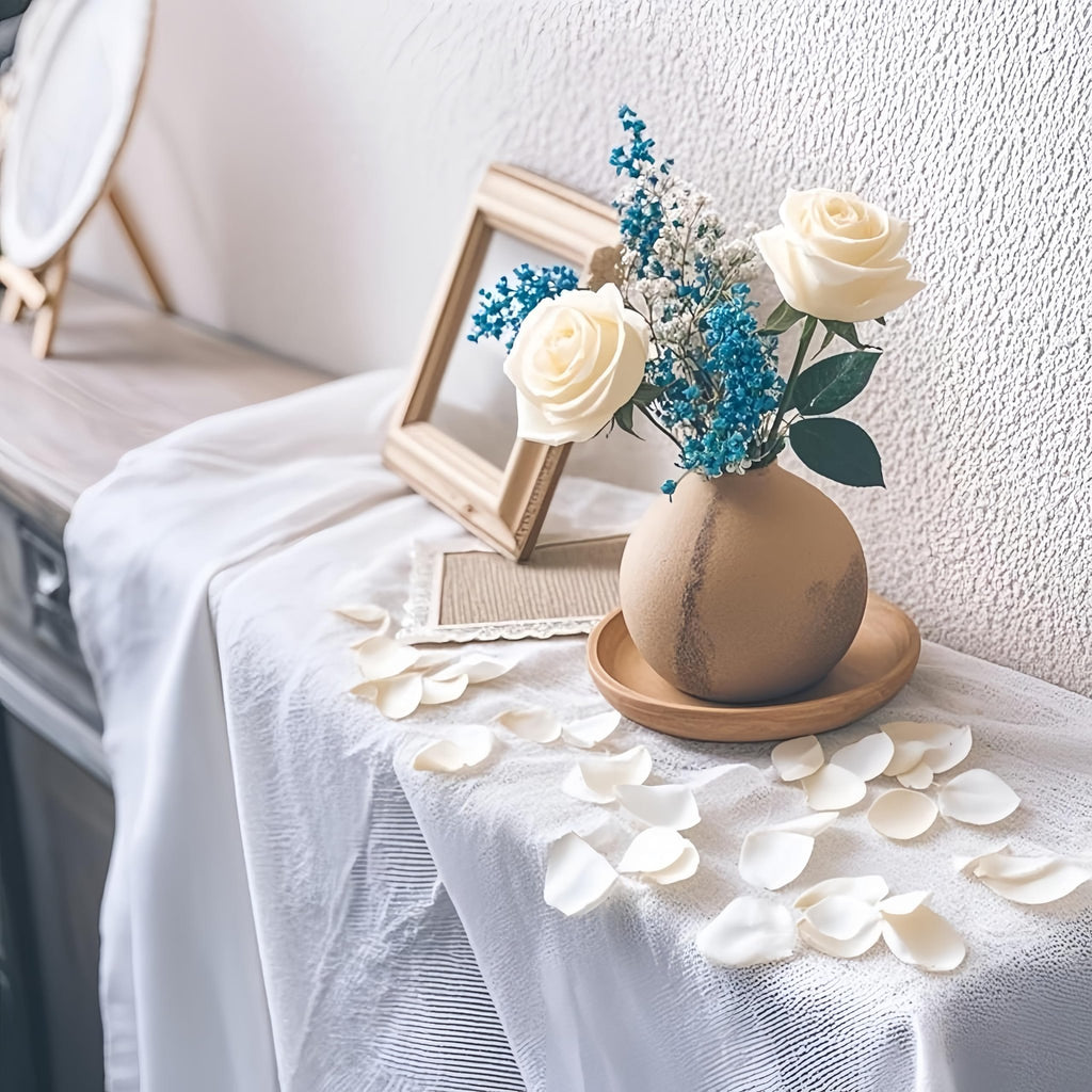 Ivory artificial rose petals scattered around a vase with white roses and blue filler flowers on a table.