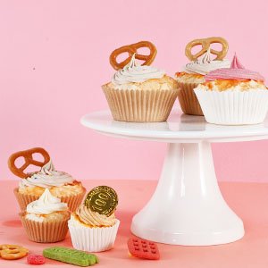 Cupcakes in natural paper liners on a white cake stand, some topped with pretzels and frosting, against a pink background.