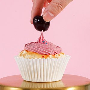 Hand placing cherry on top of frosted cupcake in white paper liner on gold stand