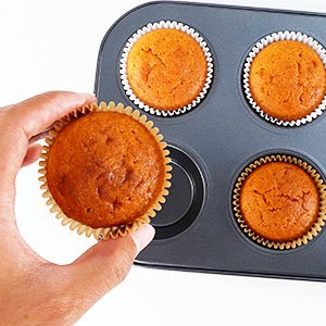 Hand holding a baked cupcake in a brown paper liner, next to a muffin tin with three more cupcakes.