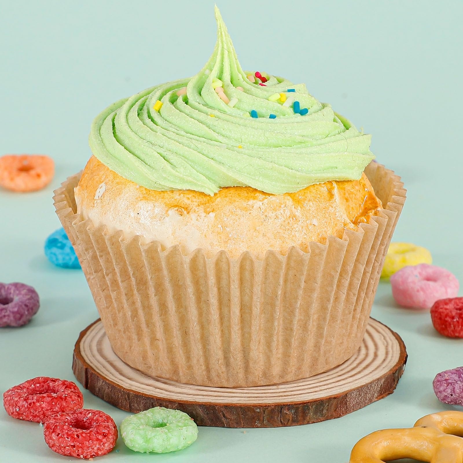 Cupcake with green frosting and sprinkles in a brown paper liner, surrounded by colorful cereal loops on a light blue background.