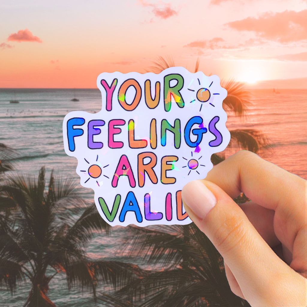 Hand holding a holographic sticker with colorful text that reads 'YOUR FEELINGS ARE VALID' against a sunset beach background.
