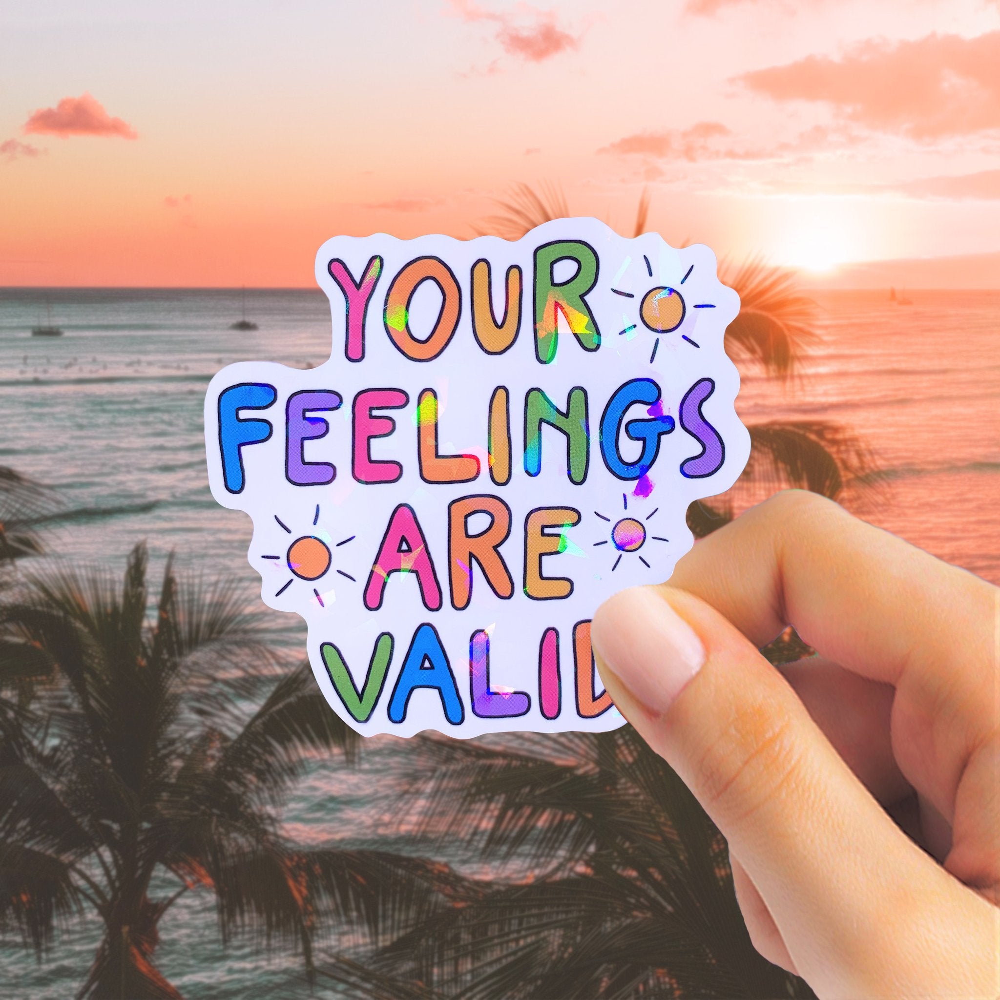 Hand holding a holographic sticker with colorful text that reads 'YOUR FEELINGS ARE VALID' against a sunset beach background.