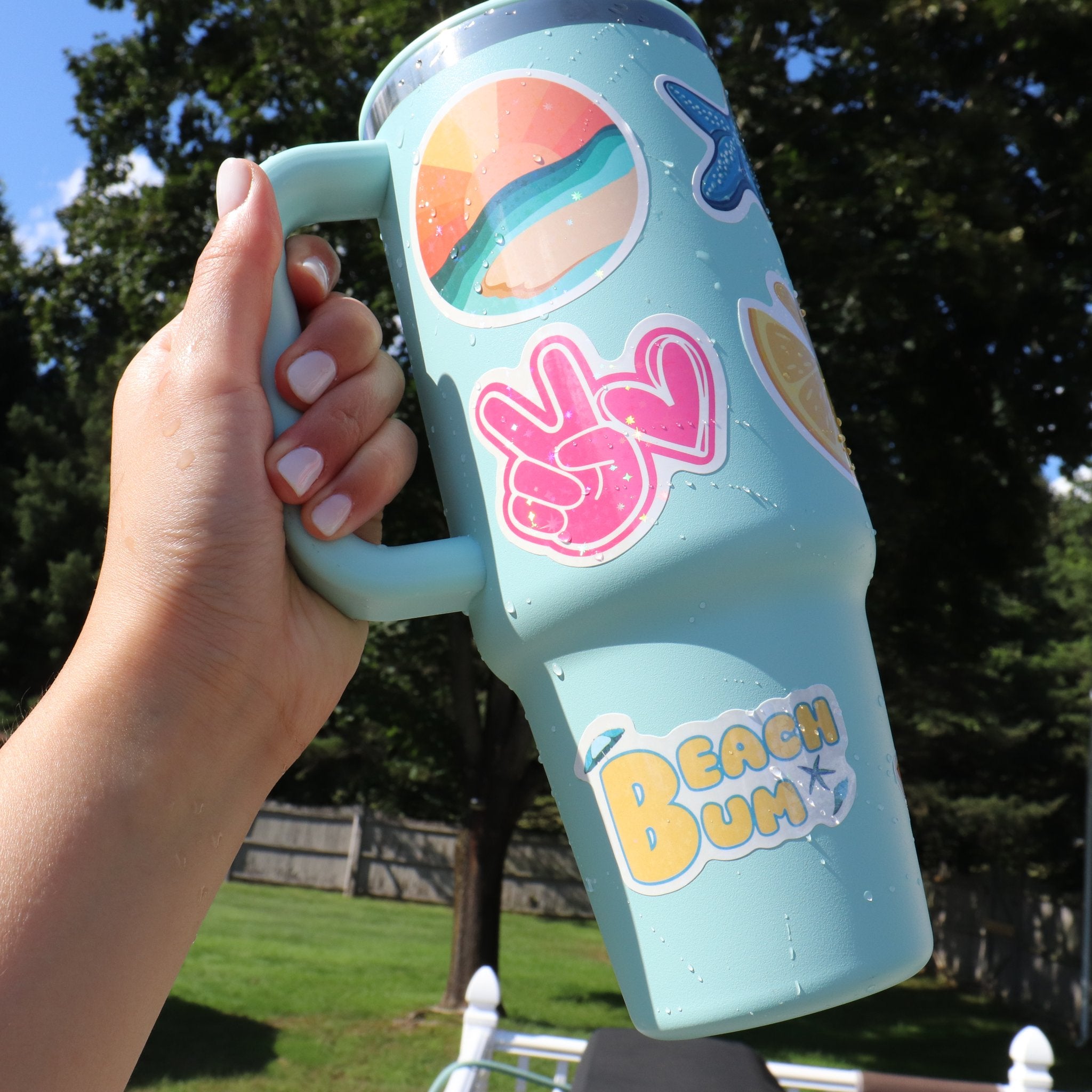 Hand holding a light blue tumbler decorated with holographic stickers including a sunset, peace sign, and "Beach Bum" text.