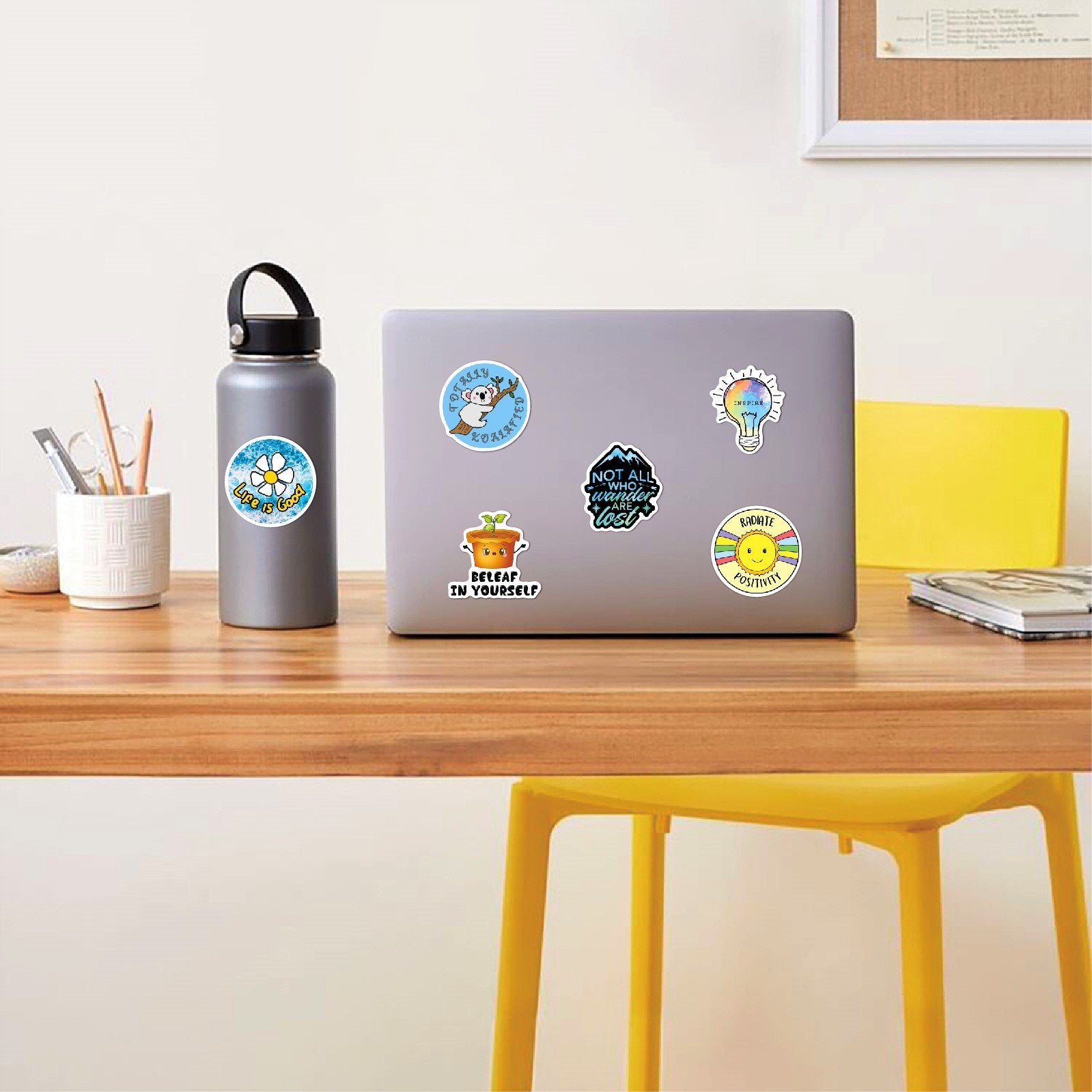 Holographic affirmation stickers applied to a laptop and water bottle on a wooden desk.
