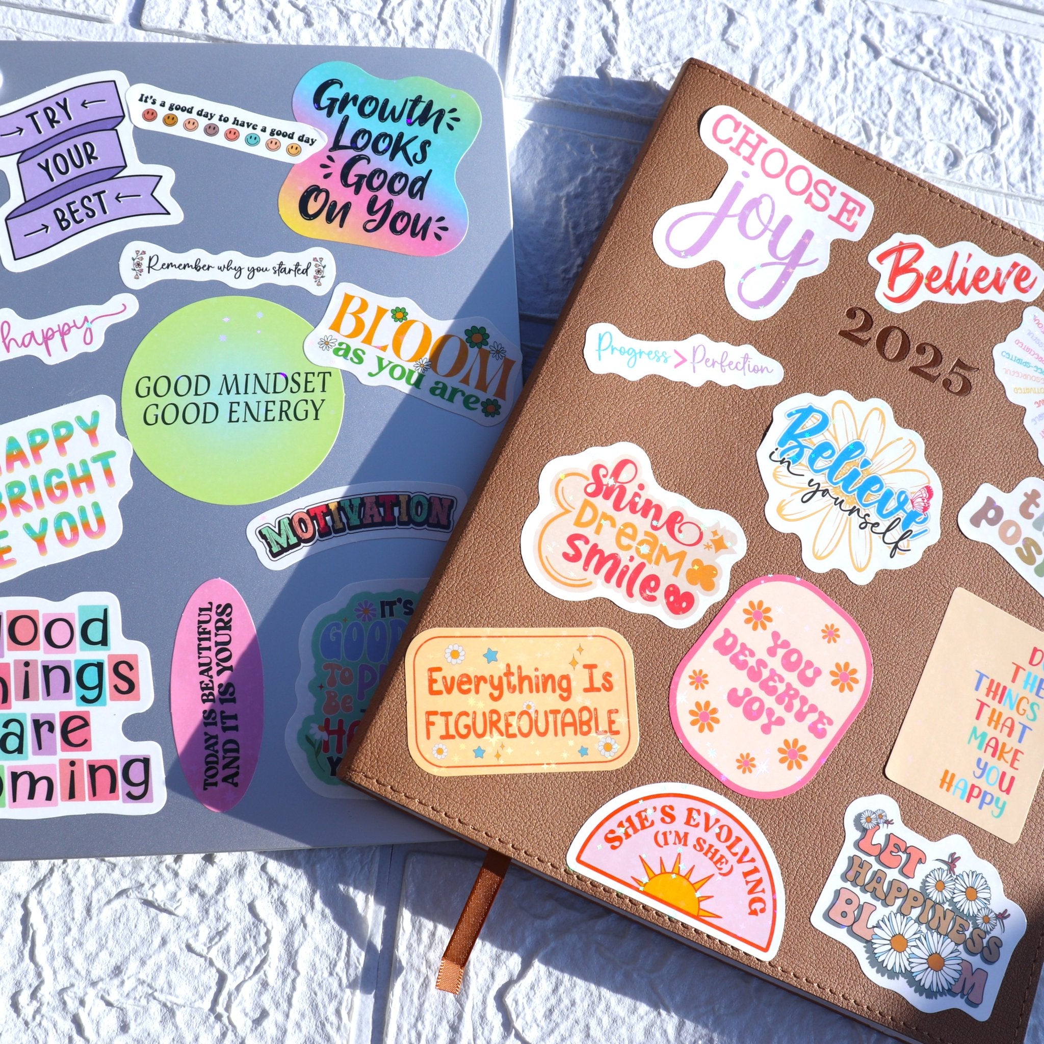 Holographic affirmation stickers applied to a laptop and a brown journal, featuring motivational quotes and colorful designs.