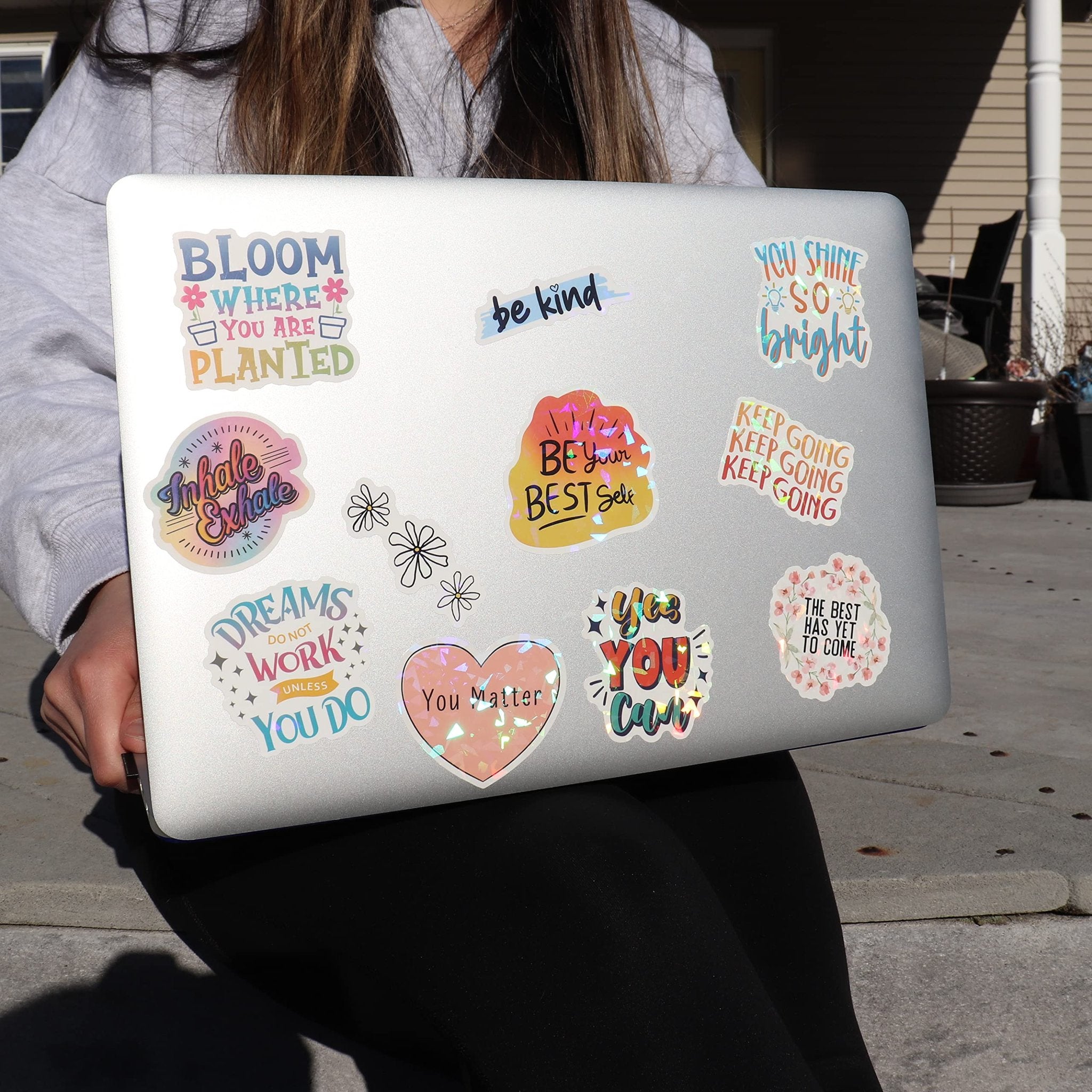 Holographic affirmation stickers applied to a laptop by a person wearing a grey hoodie.