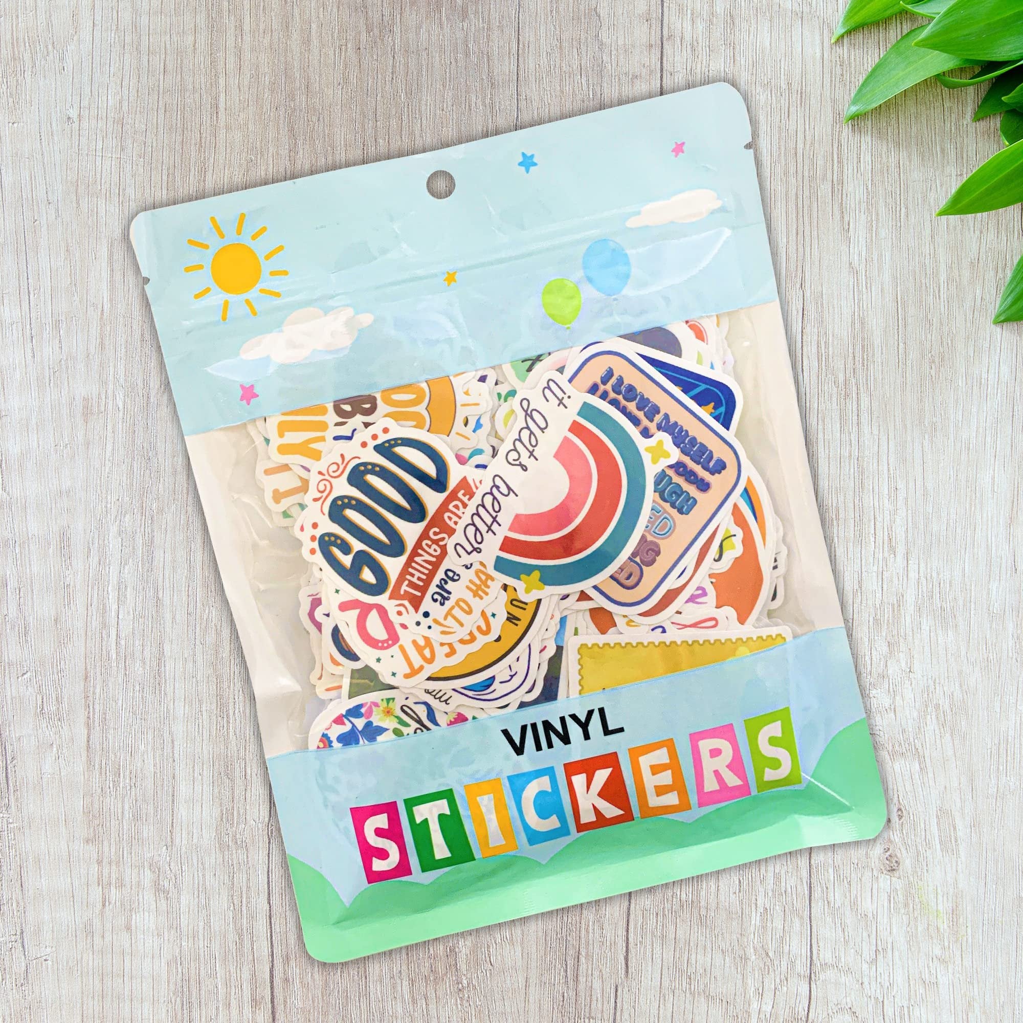 Bag of holographic affirmation stickers with motivational quotes, on a wooden background with green leaves.