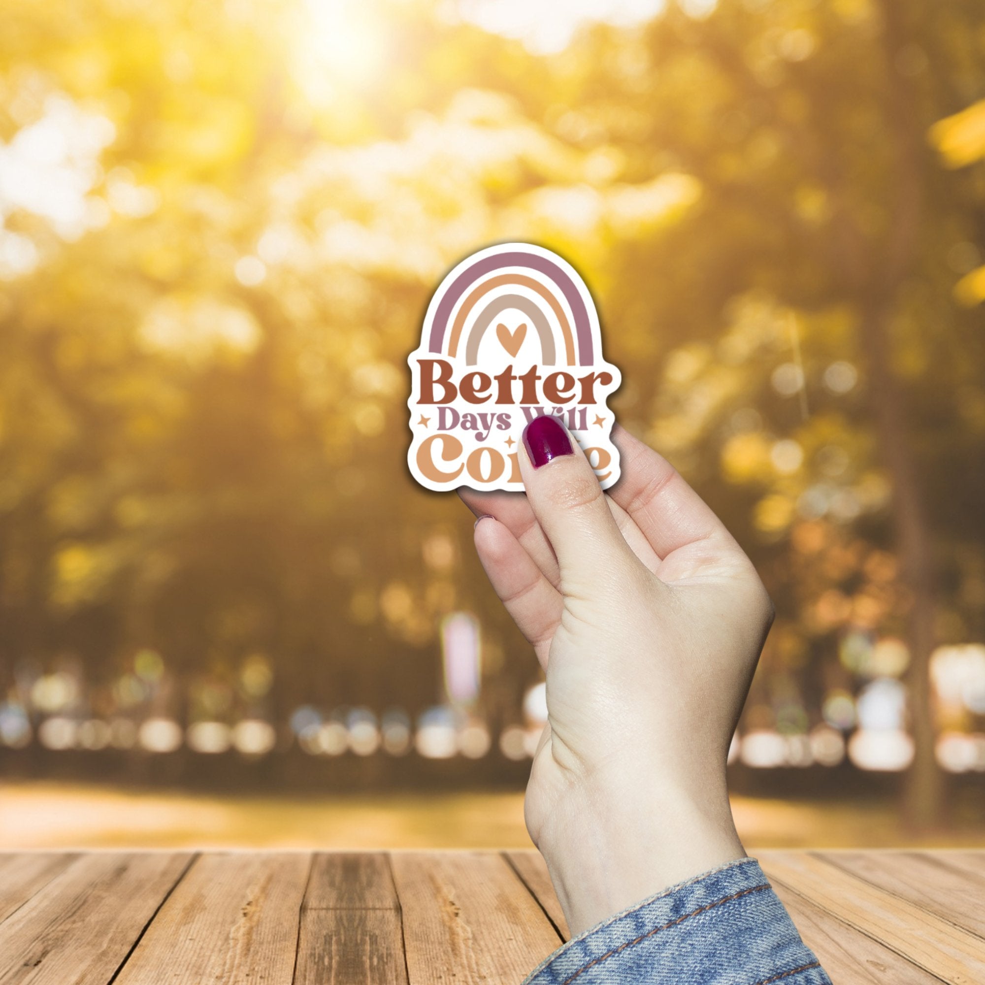 Hand holding a boho-style vinyl sticker with text 'Better Days Will Come' against a blurred outdoor background.