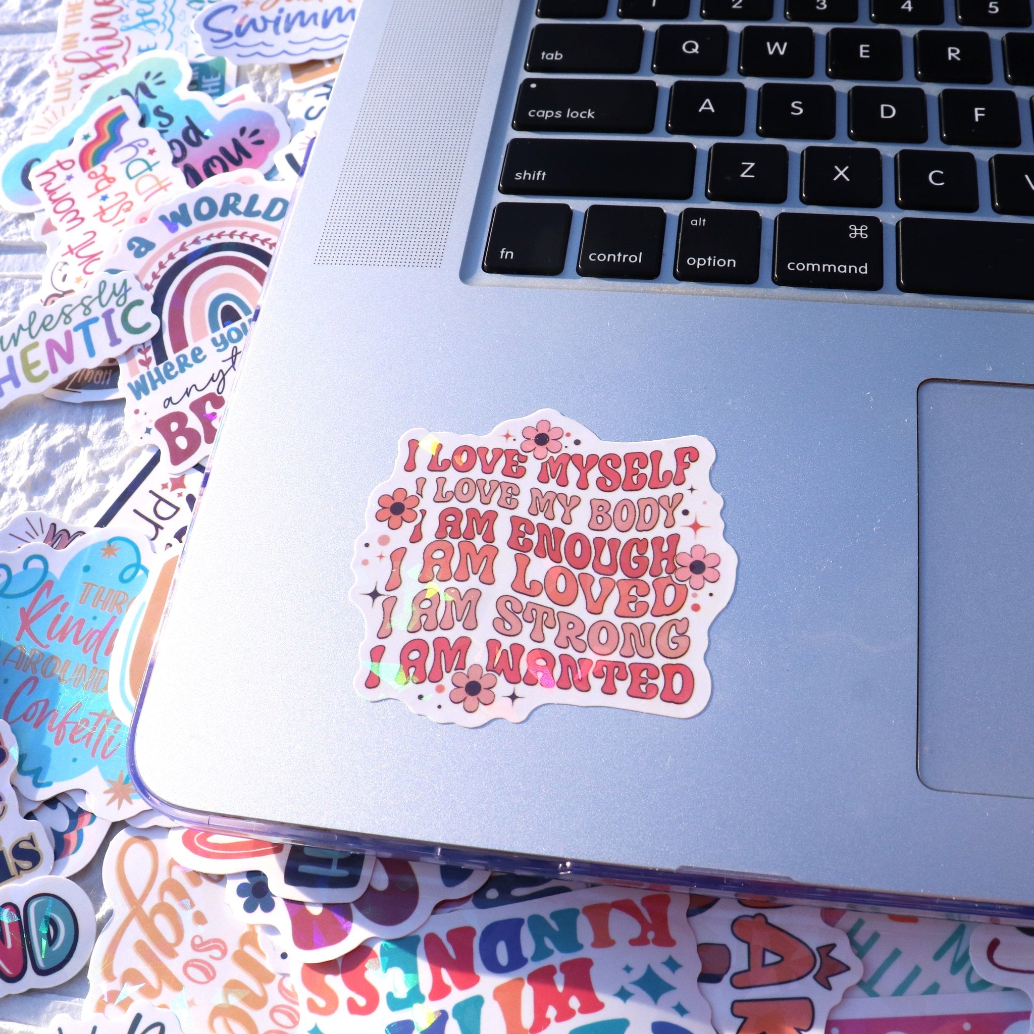 Motivational vinyl stickers with inspirational quotes scattered around a laptop keyboard. One sticker reads 'I LOVE MYSELF. I LOVE MY BODY. I AM ENOUGH. I AM LOVED. I AM STRONG. I AM WANTED.'