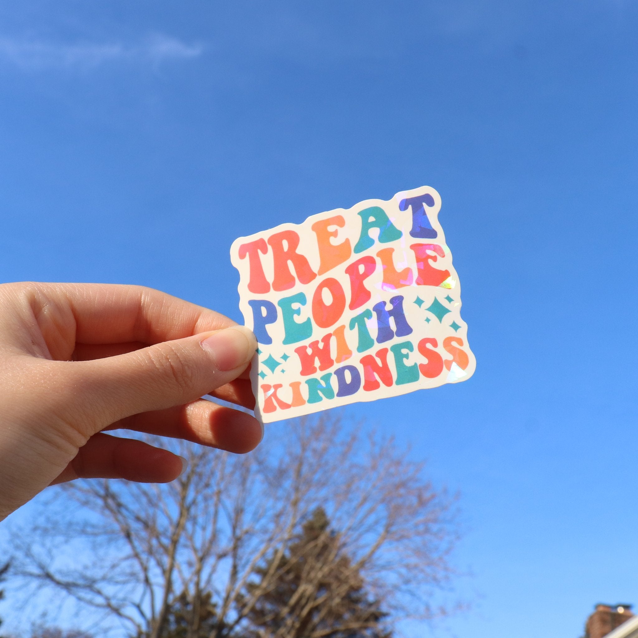 Hand holding a holographic vinyl sticker with colorful wavy text that says 'TREAT PEOPLE WITH KINDNESS' against a bright blue sky.