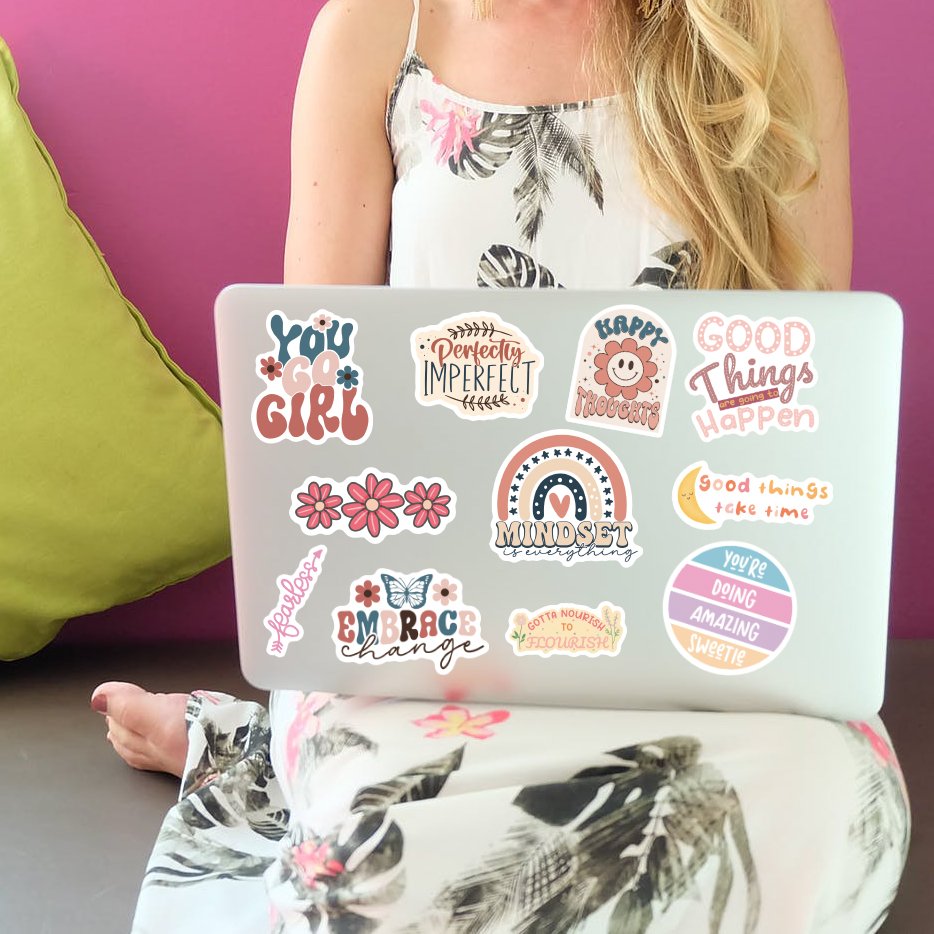Woman sitting with a laptop covered in motivational vinyl stickers. Stickers feature quotes like 'You Go Girl' and 'Good Things Happen'.