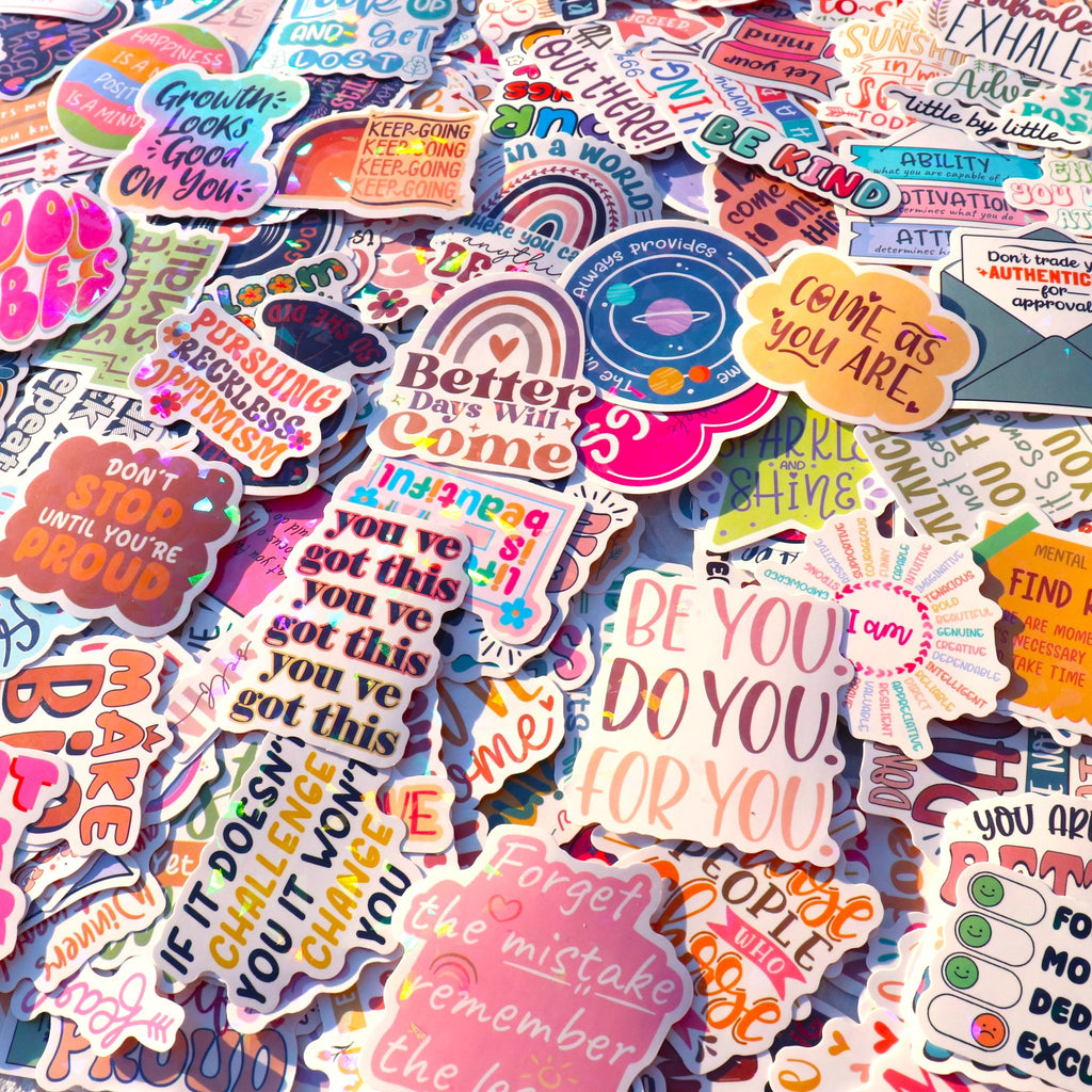 A flat lay of many holographic vinyl stickers with motivational and boho quotes, scattered across a surface.