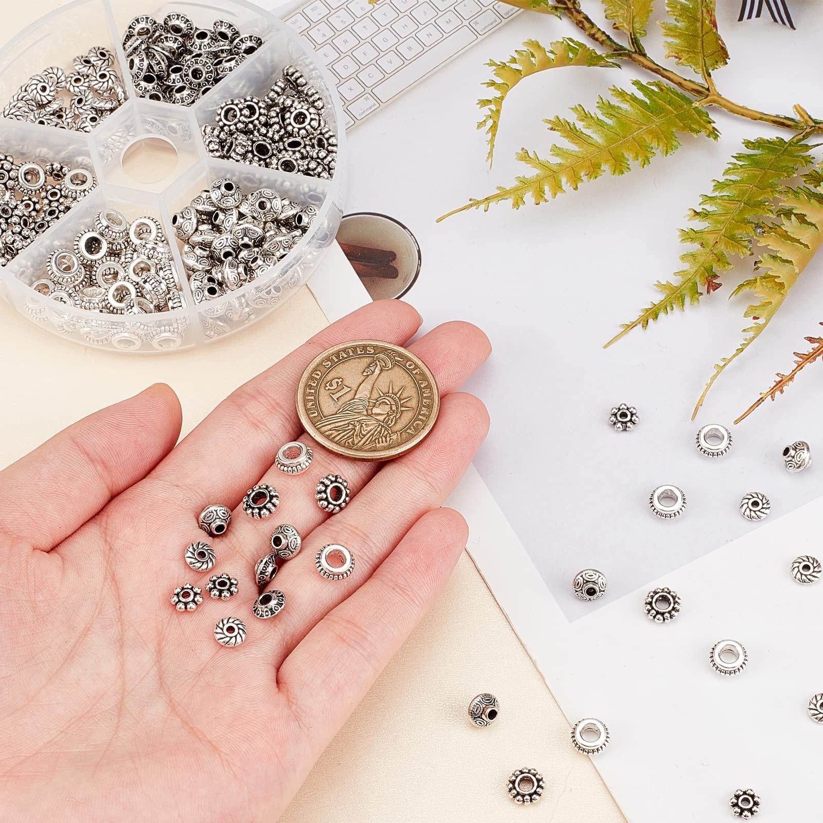 Hand holding antique silver metal spacer beads with a US dollar coin for scale, next to a compartment organizer filled with beads.