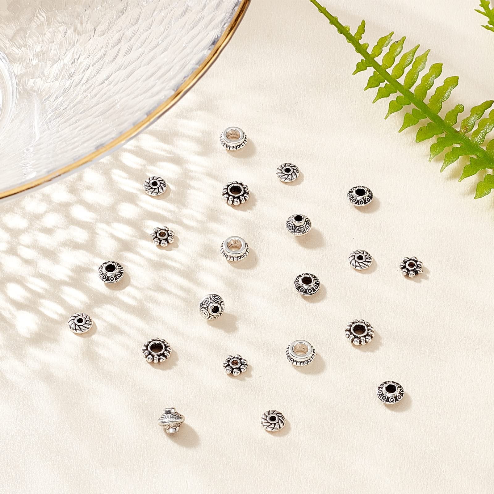 Assortment of antique silver metal spacer beads with various intricate designs scattered on a light-colored surface with shadows and a fern leaf.