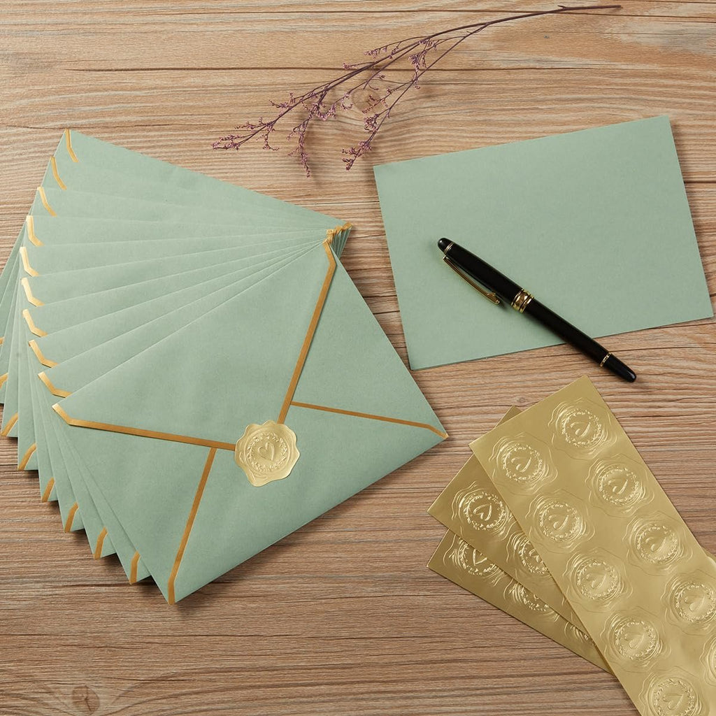 Stack of sage green envelopes with gold trim and a gold heart seal, next to a blank card and a pen, with gold heart stickers on a sheet.