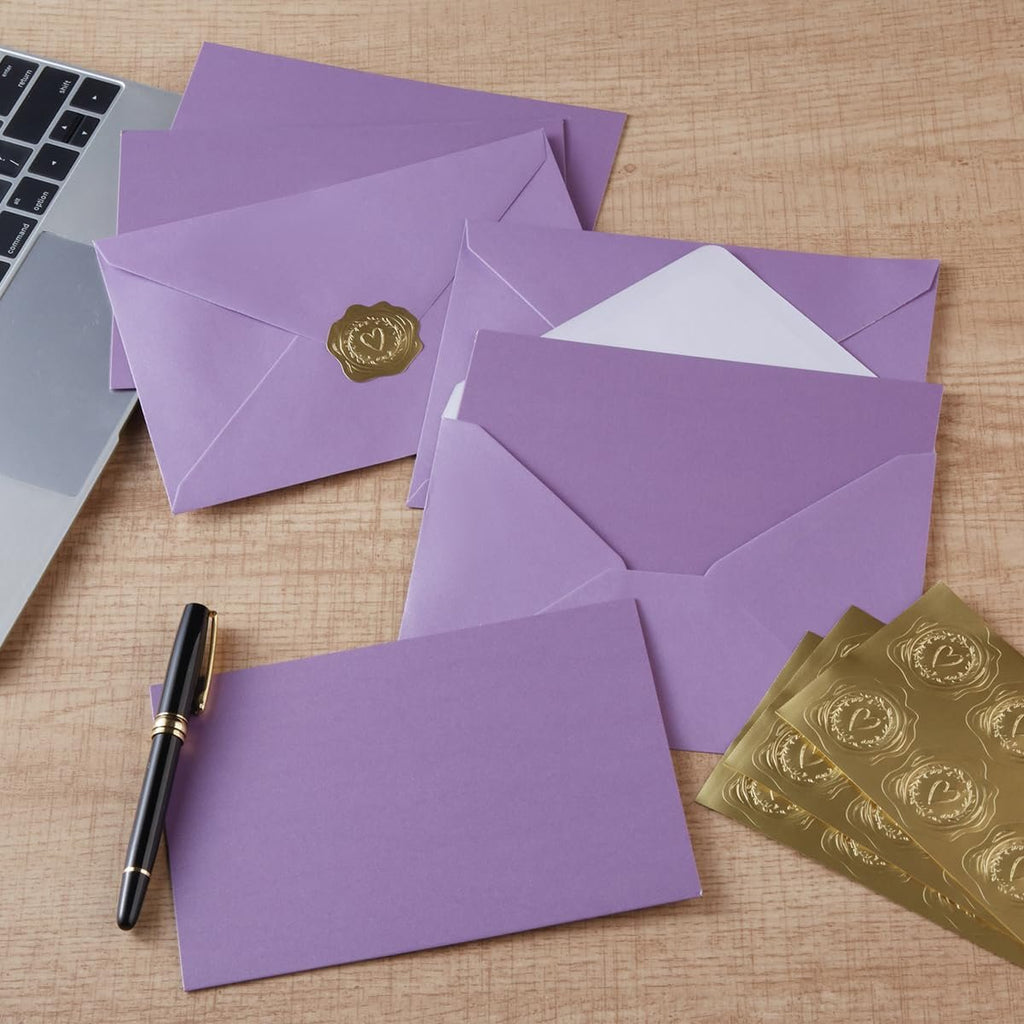 Purple envelopes and gold heart seal stickers arranged on a wooden surface next to a laptop and pen.