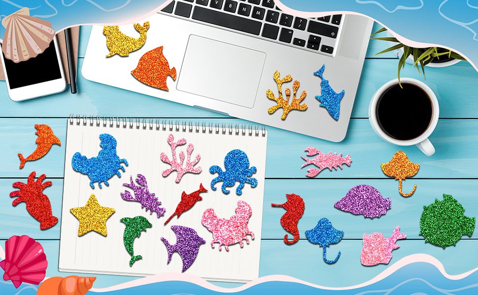 Glitter sea animal foam stickers scattered on a desk with a laptop, notebook, and coffee cup.