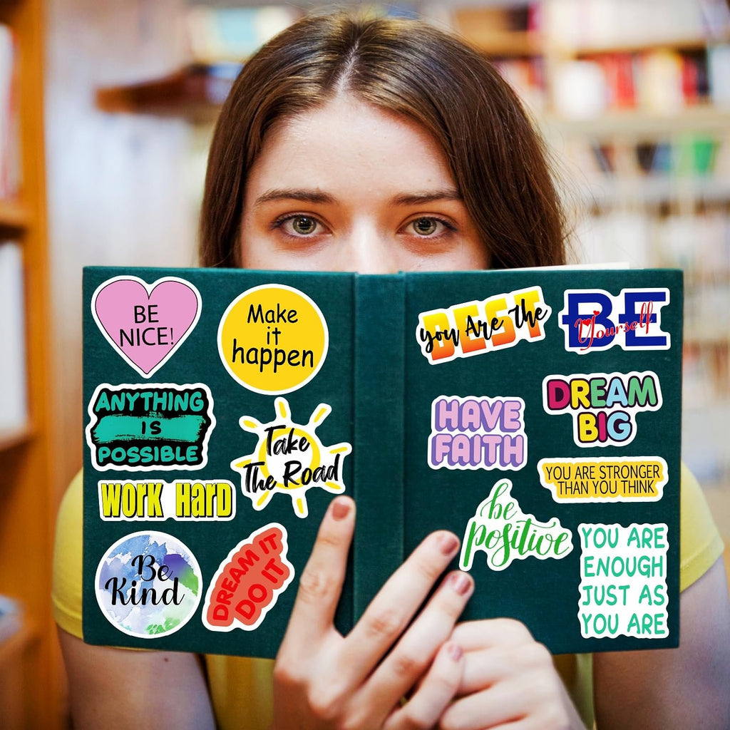 Young woman holding a book covered in inspirational quote stickers. Stickers include 'Be Nice', 'Make it Happen', 'Work Hard', 'Dream Big', and 'Be Kind'.