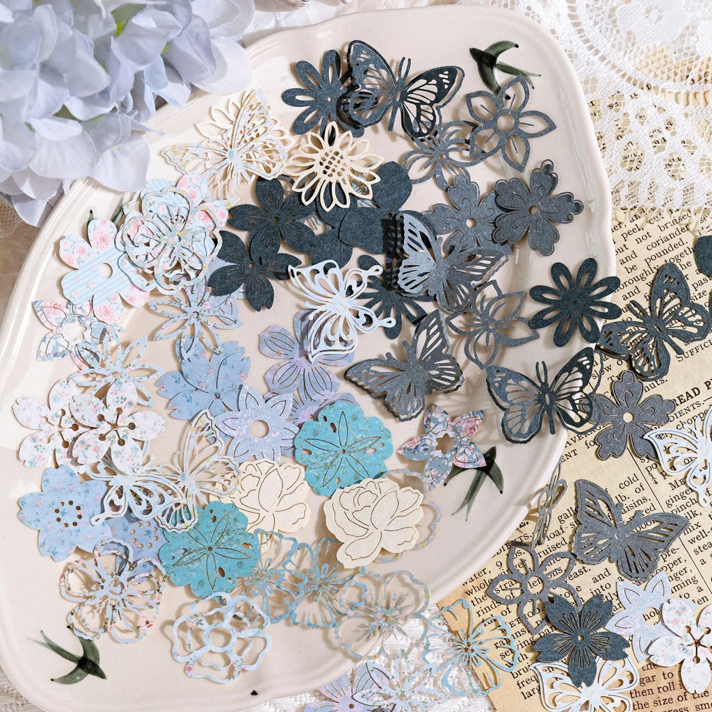 Collage paper cutouts of flowers and butterflies in various pastel and dark gray colors scattered on a decorative plate.