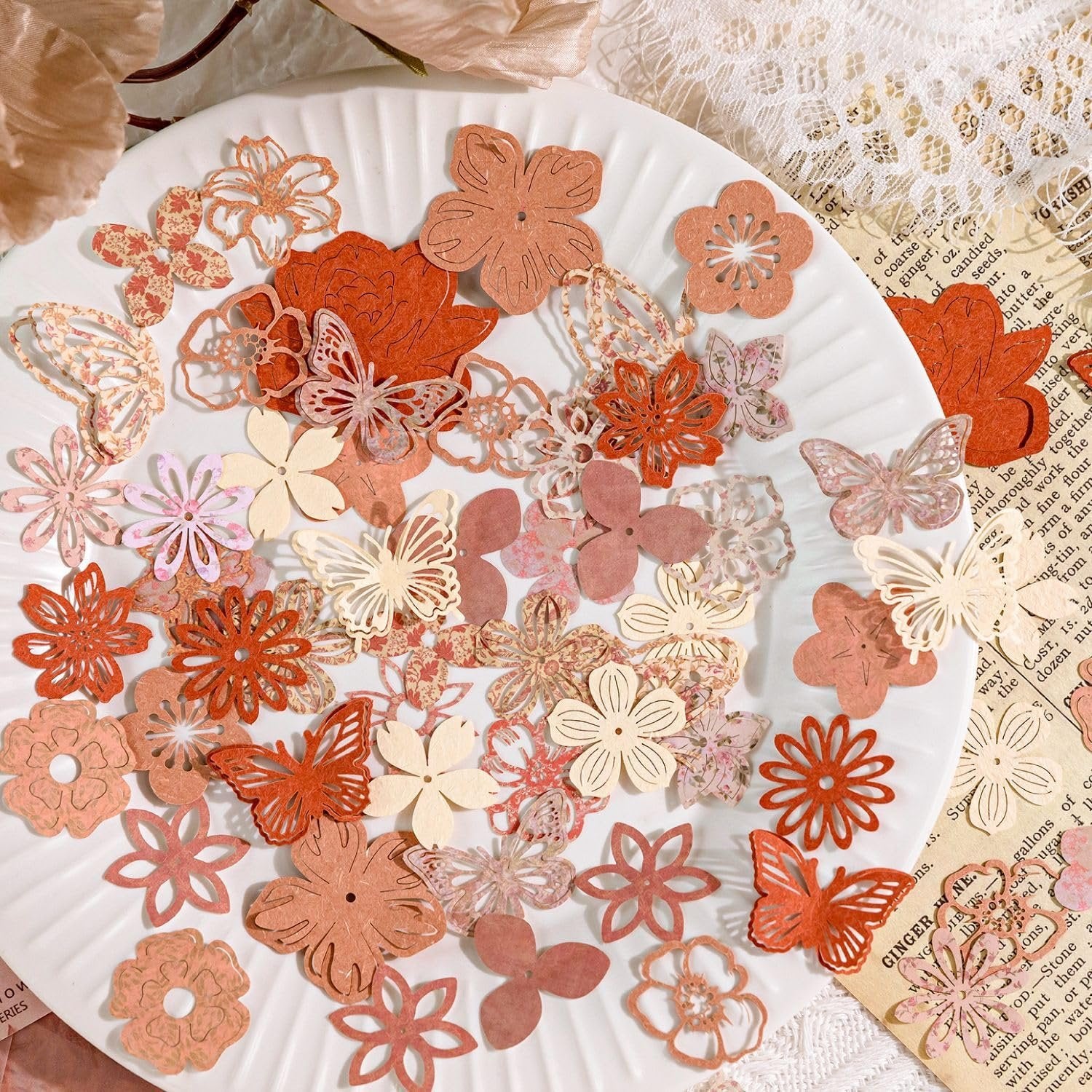 Collage paper cutouts of flowers and butterflies scattered on a white plate, with vintage paper and lace in the background.