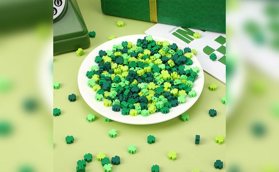 Pile of green shamrock polymer clay beads in four shades on a white plate, with scattered beads around.