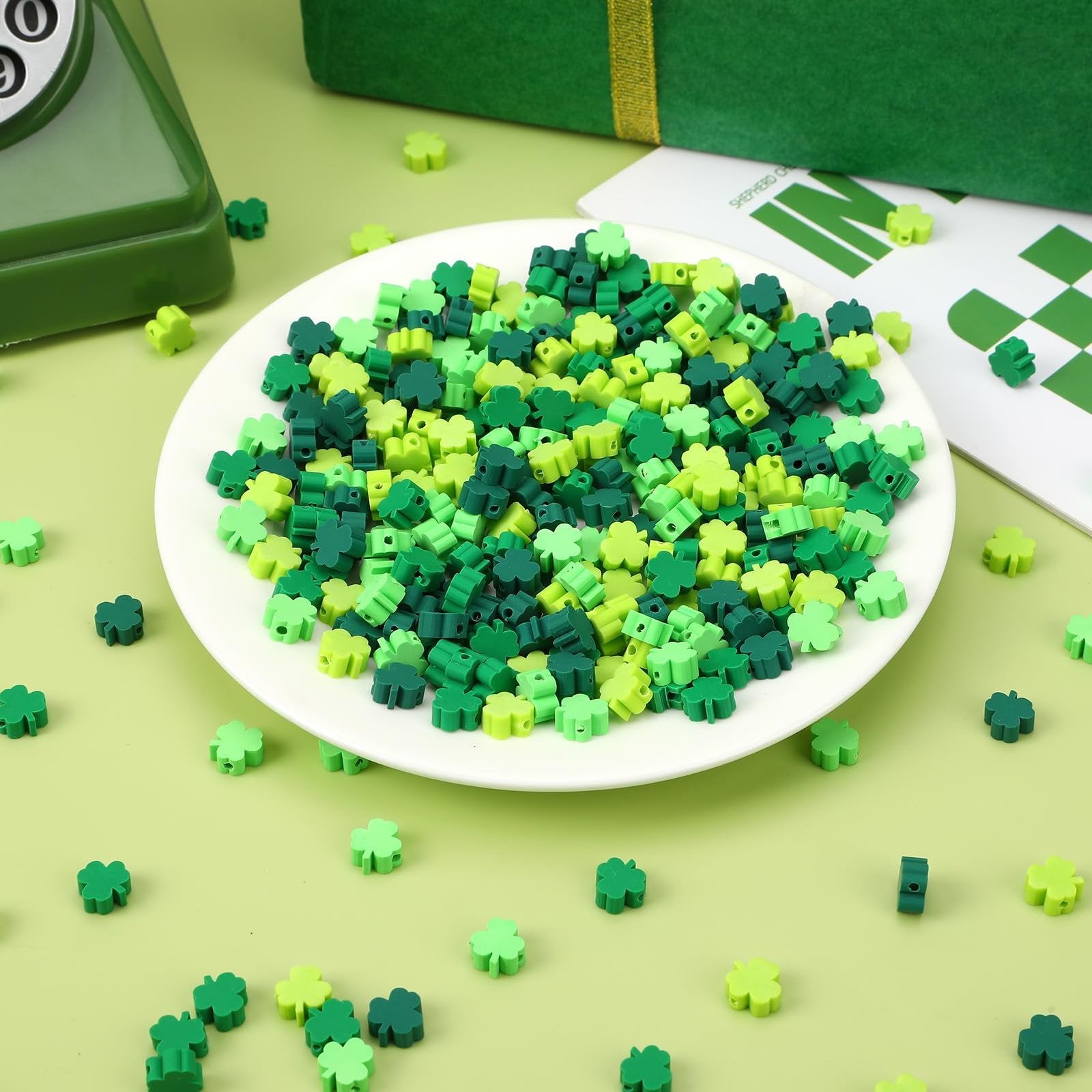 Pile of green shamrock polymer clay beads in four shades on a white plate, with scattered beads on a light green surface.