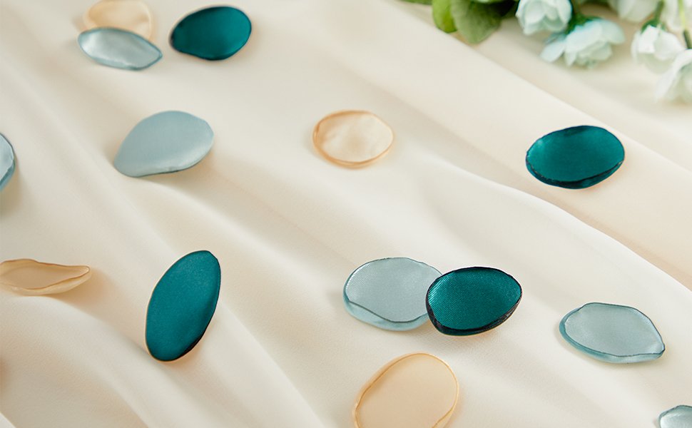 Scattered silk rose petals in champagne, light blue, and teal on a draped cream fabric with greenery in the background.