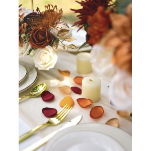 Terracotta satin rose petals scattered on a table with candles and floral arrangements for a wedding setting.